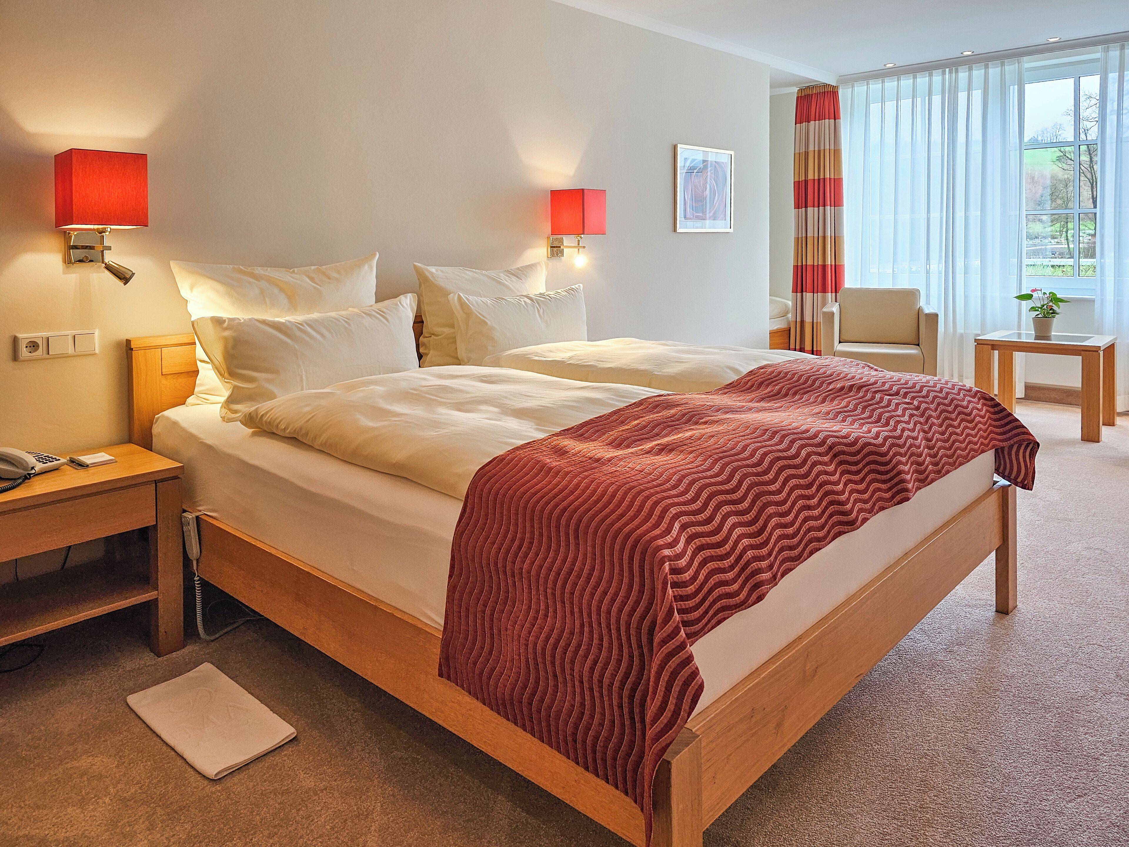 Quarto de hotel com uma cama grande, duas luminárias de cabeceira, uma mesa pequena com uma planta e vista para o exterior através de uma janela com cortinas.