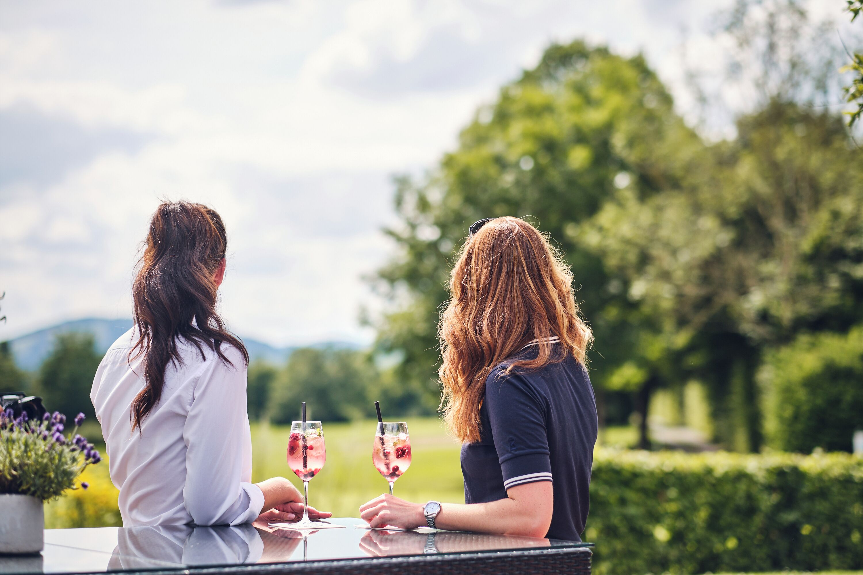 Two people sitting at a table outdoors with drinks, surrounded by greenery and a clear sky.