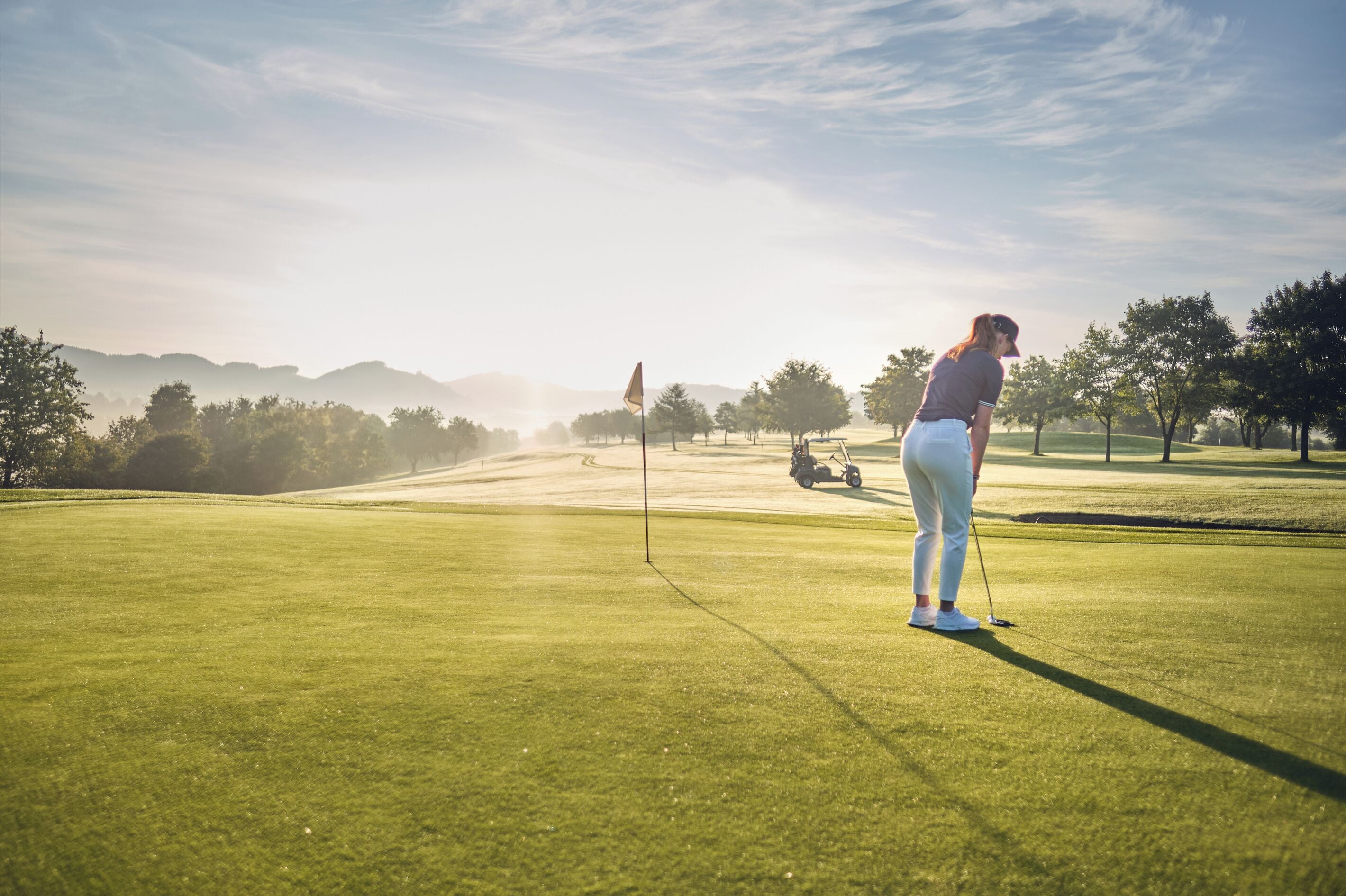 A golf course with a flagstick, a golf cart, and a golfer in the distance.