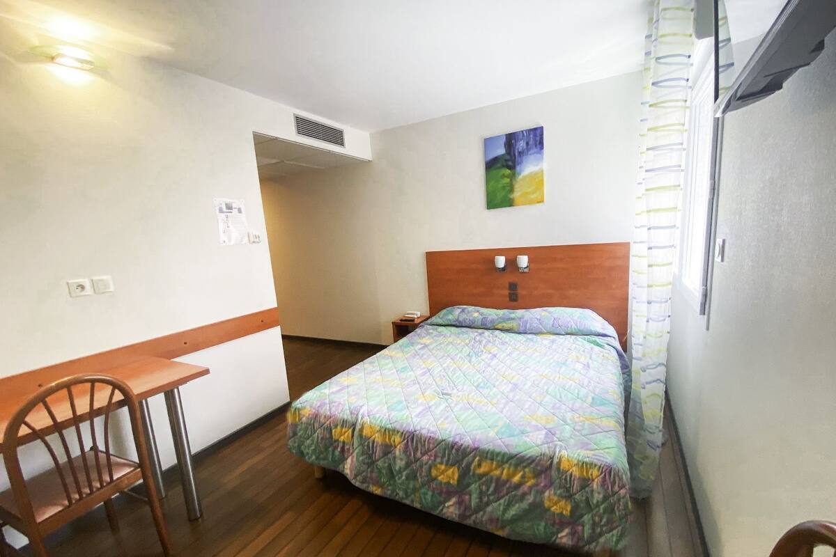 Una cama individual con un edredón colorido, un escritorio de madera y una silla en una habitación con paredes blancas y suelo de madera.