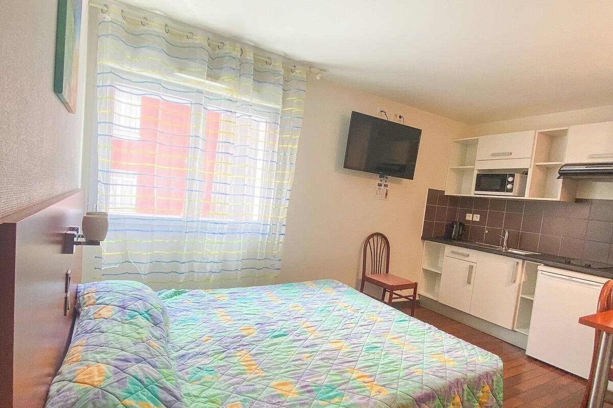 Un dormitorio con cama, televisor montado en la pared, ventana con cortinas y una kitchenette con microondas y armarios.