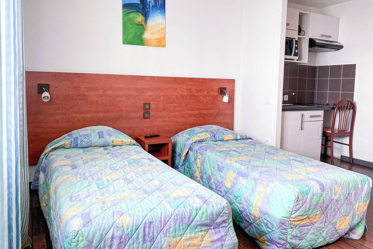 Una habitación con dos camas, cabecero de madera, un cuadro en la pared, una mesita y una kitchenette.