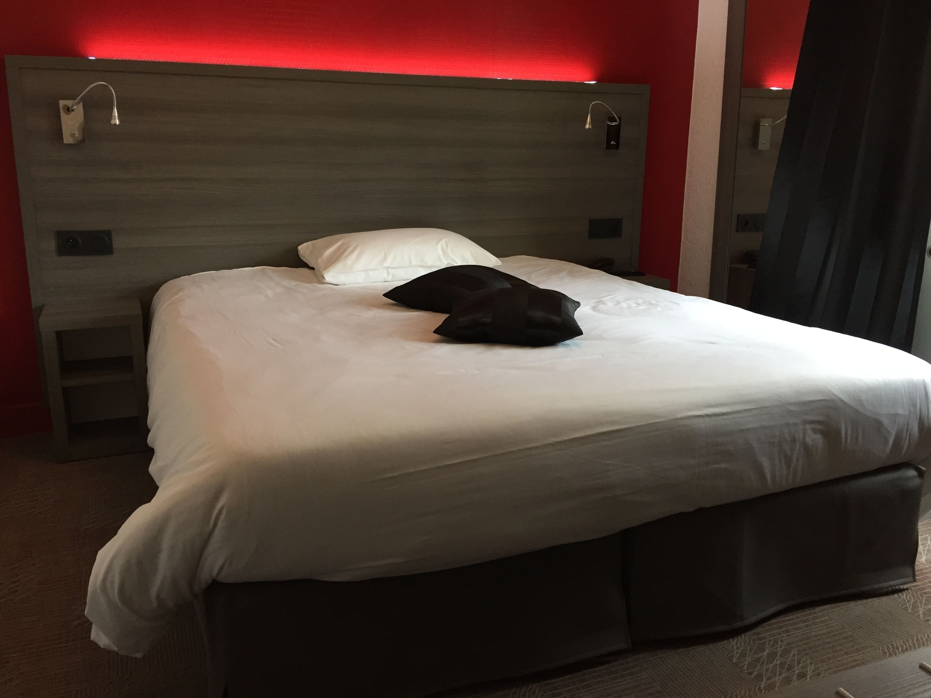 Una cama bien hecha con ropa blanca y una almohada negra, un cabecero de madera y una pared roja.