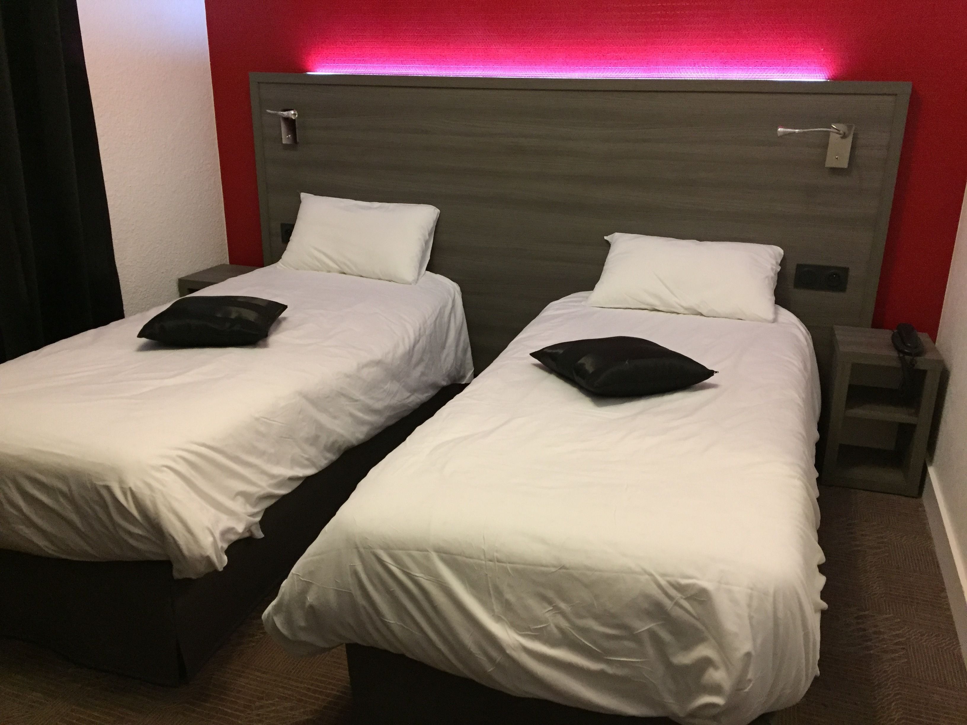 Dos camas individuales con ropa de cama blanca y almohadas negras, un cabecero de madera y una pared de acento roja.