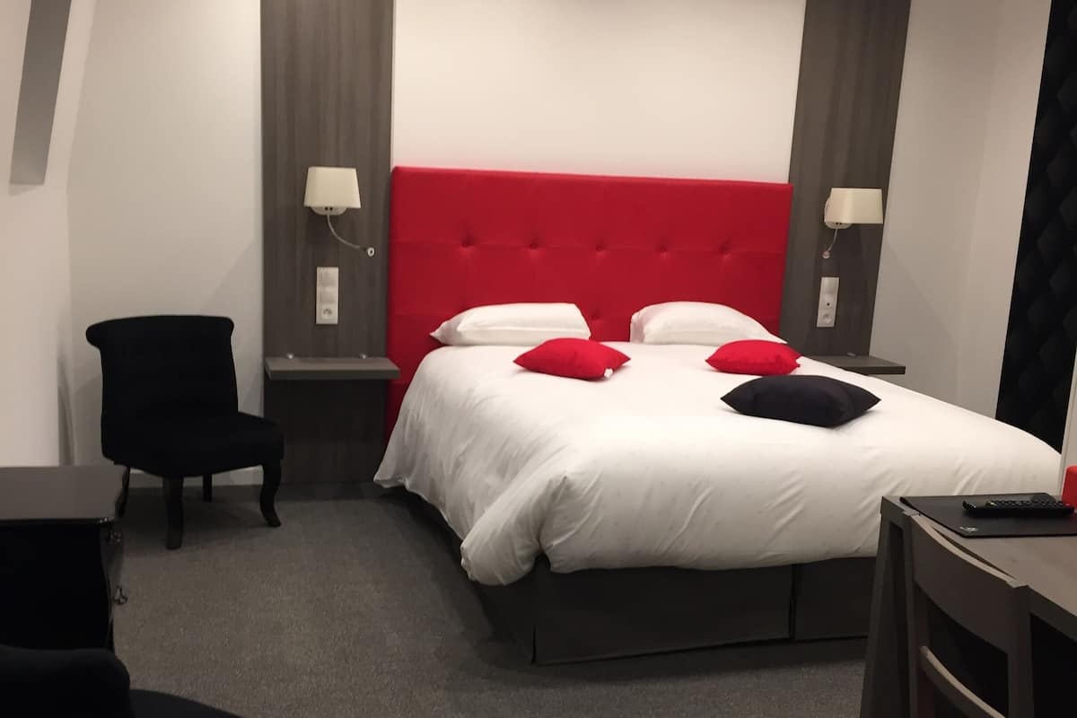 A hotel room with a red headboard, a bed with white and red pillows, a desk with a chair, and two bedside lamps.