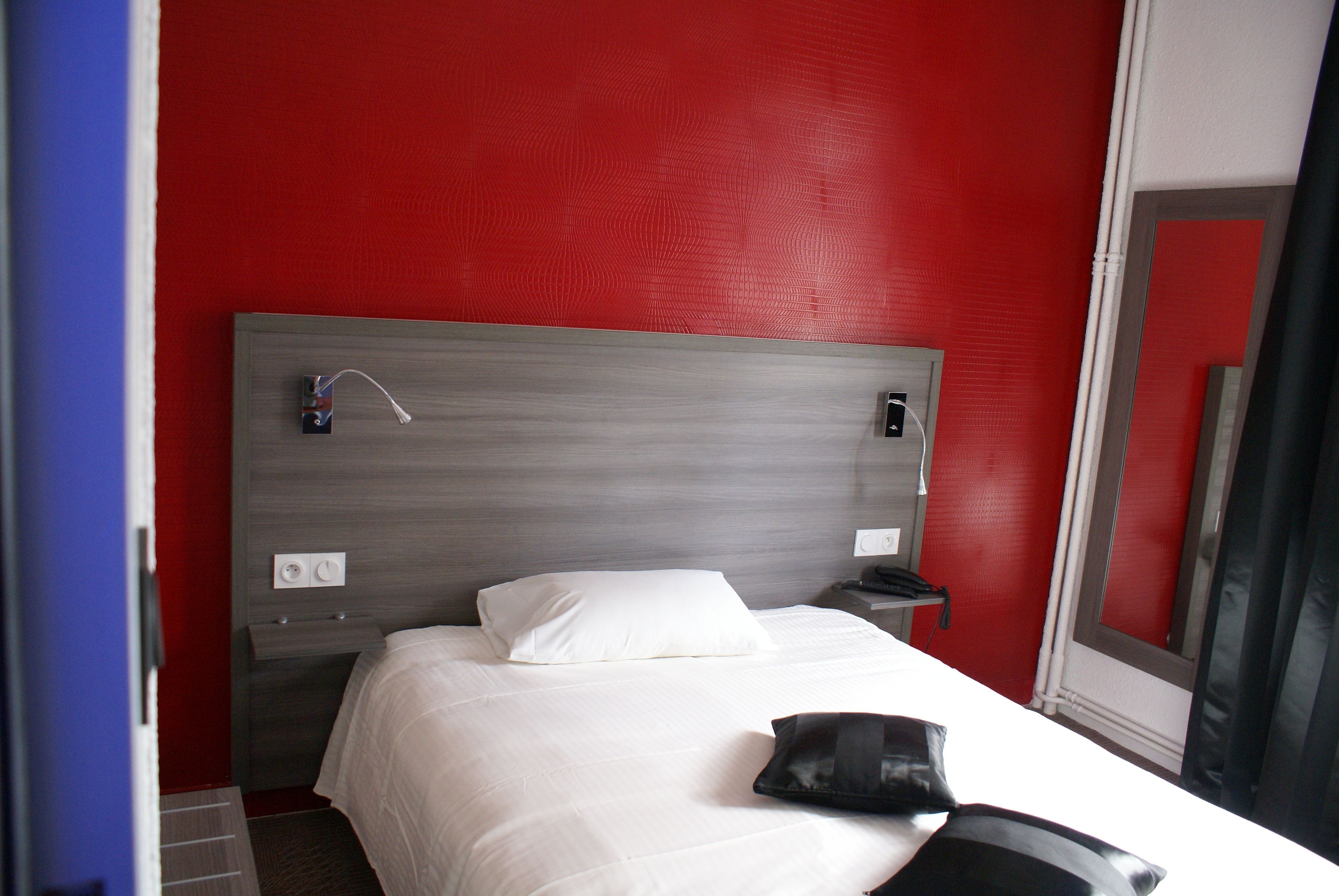 A hotel room with a red wall, a grey headboard, a white bed, a bedside table with a phone, and a mirror.