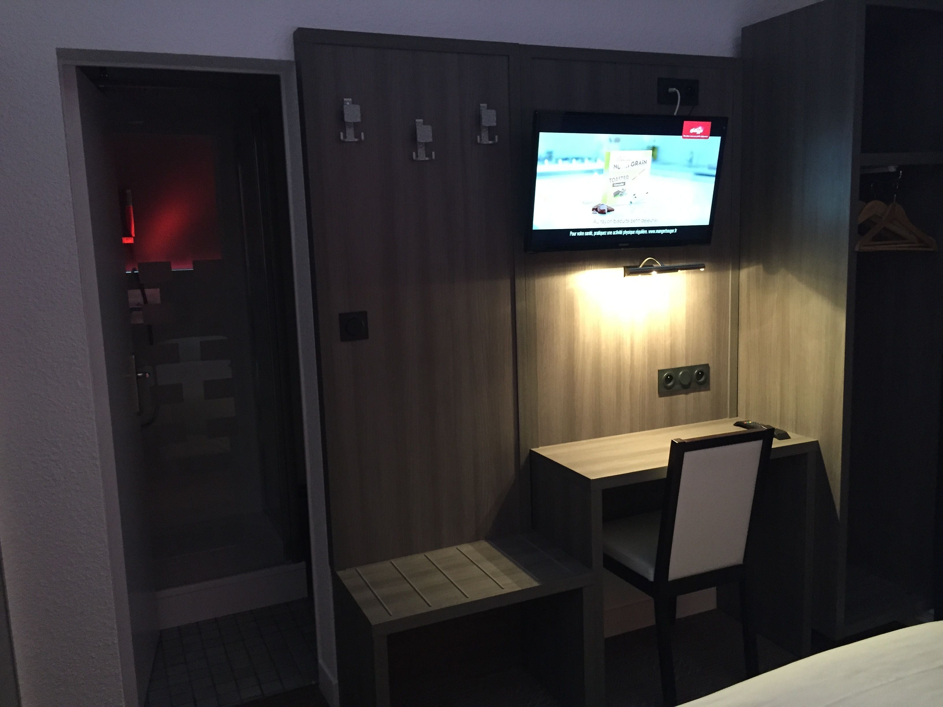 A hotel room with a wooden wardrobe, a desk with a chair, and a television mounted on the wall.