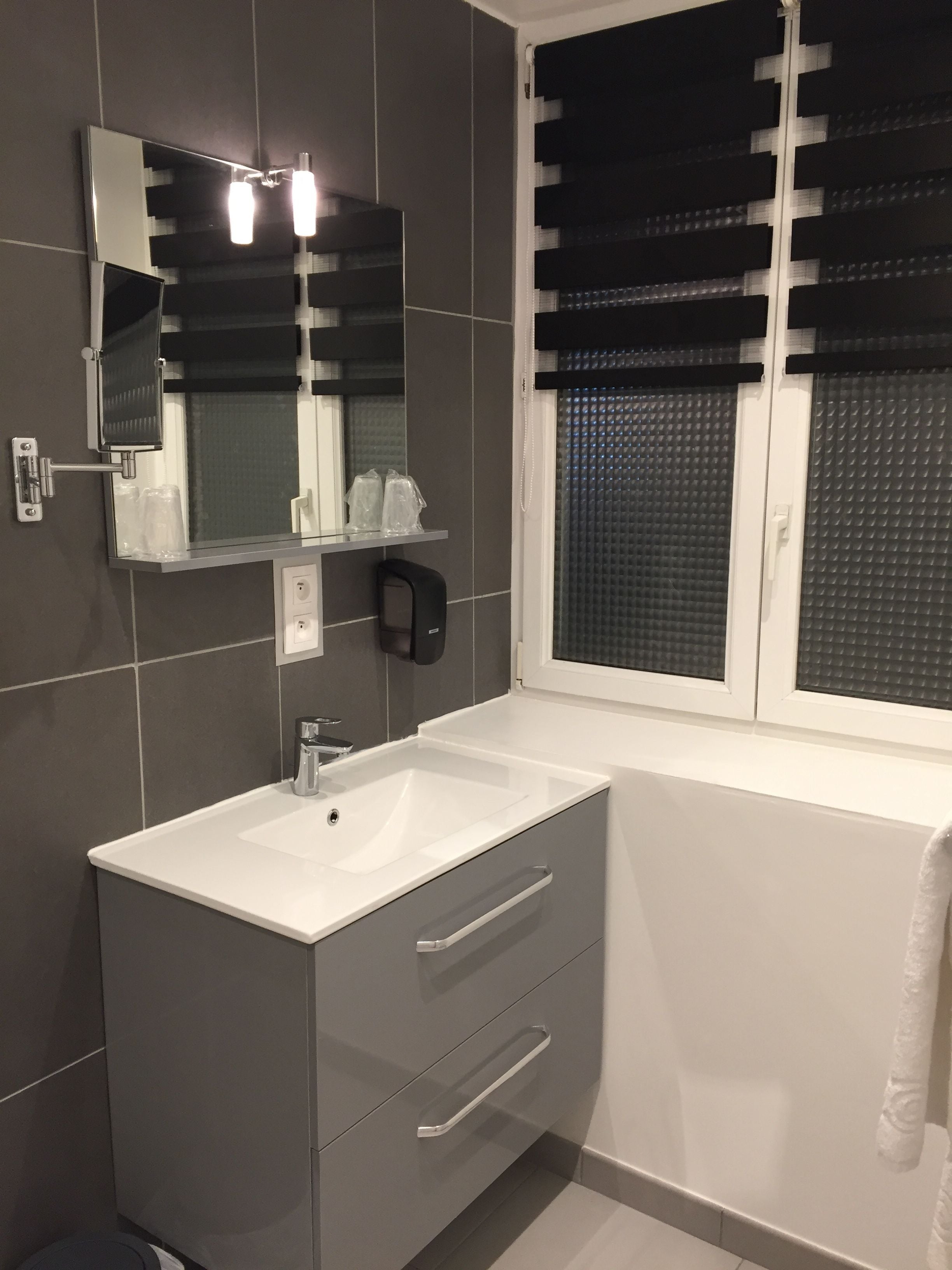 A modern bathroom with a white sink, a mirror, and a window with black blinds.