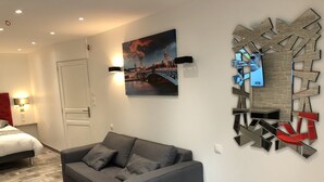 Una habitación de hotel moderna con un sofá gris, un cuadro de un paisaje urbano, una original pieza de arte con espejos y una pequeña mesa redonda.