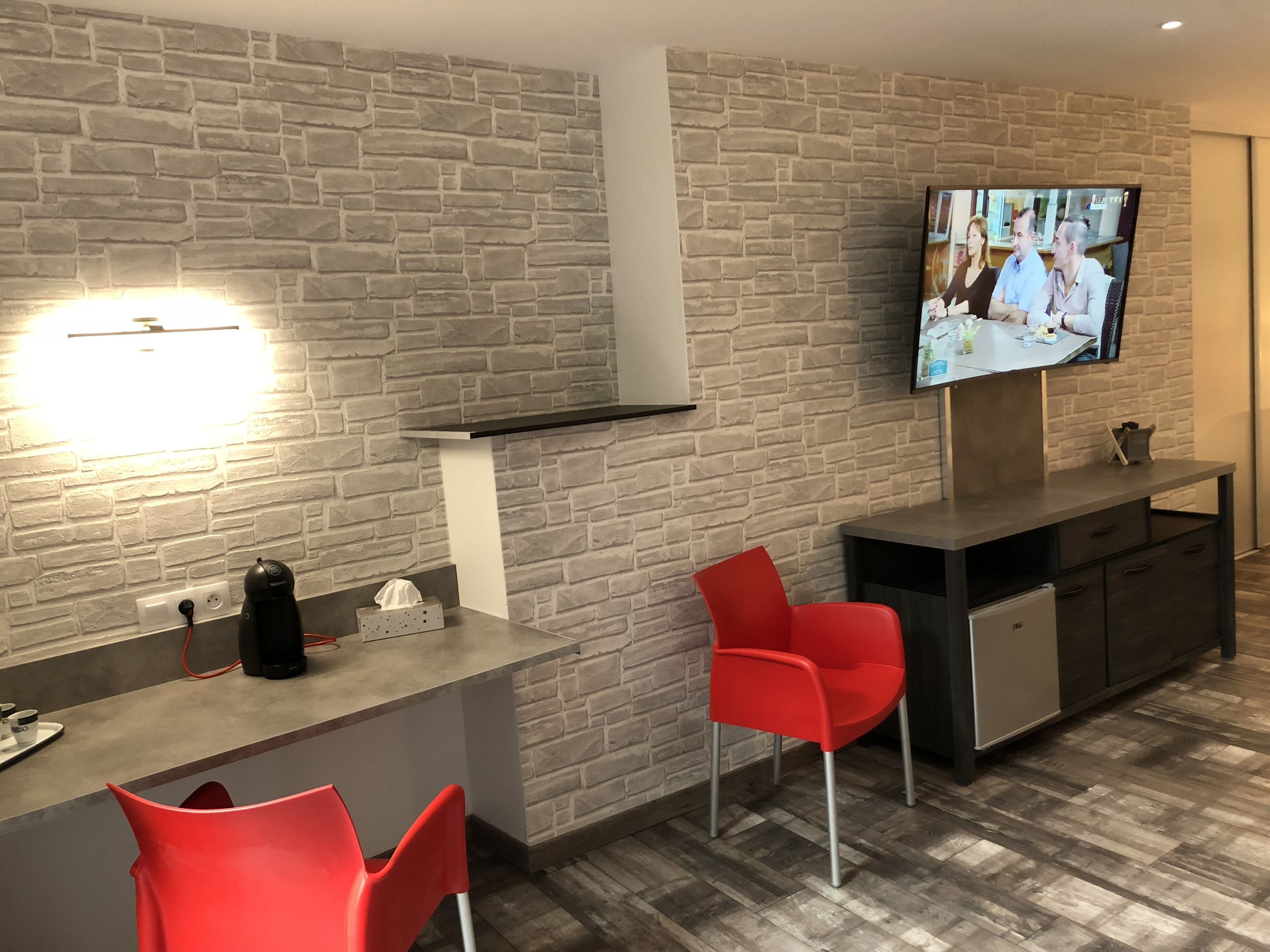 Une cuisine moderne avec un mur de pierres apparentes derrière l’évier, un téléviseur à écran plat fixé au-dessus d’un plan de travail foncé, et deux chaises rouges.
