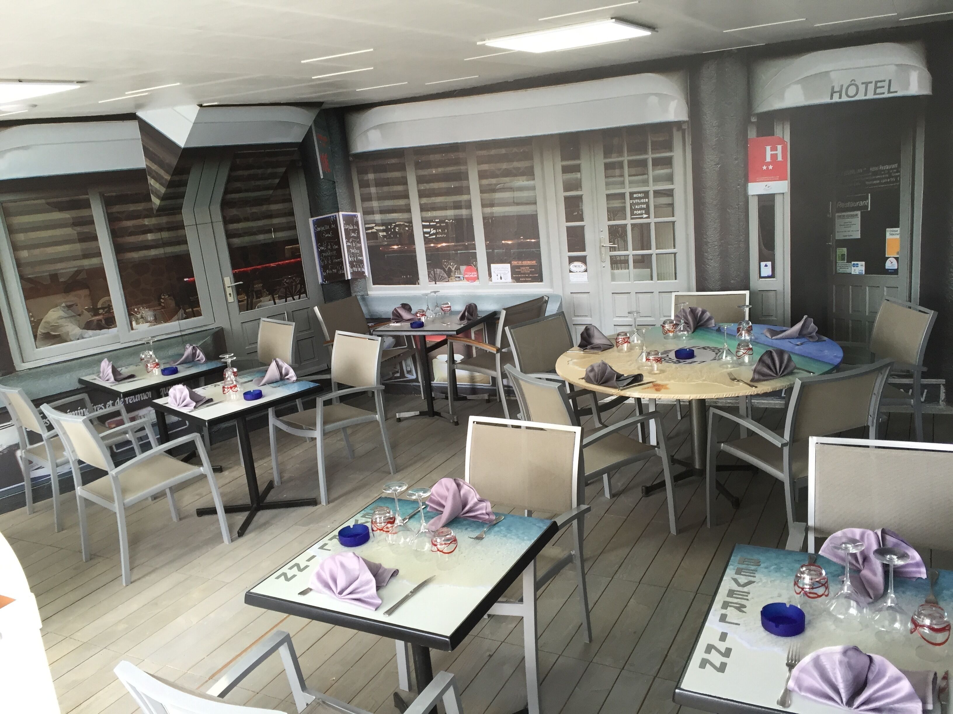 A restaurant with tables set for dining, a visible 'Hôtel' sign, and a view through large windows.