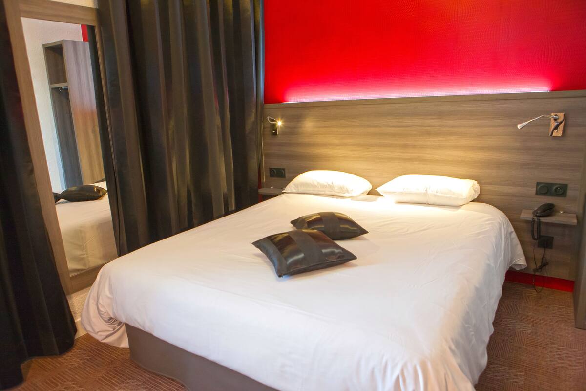A hotel room with a bed, pillows, a bedside table with a phone, and a red wall.