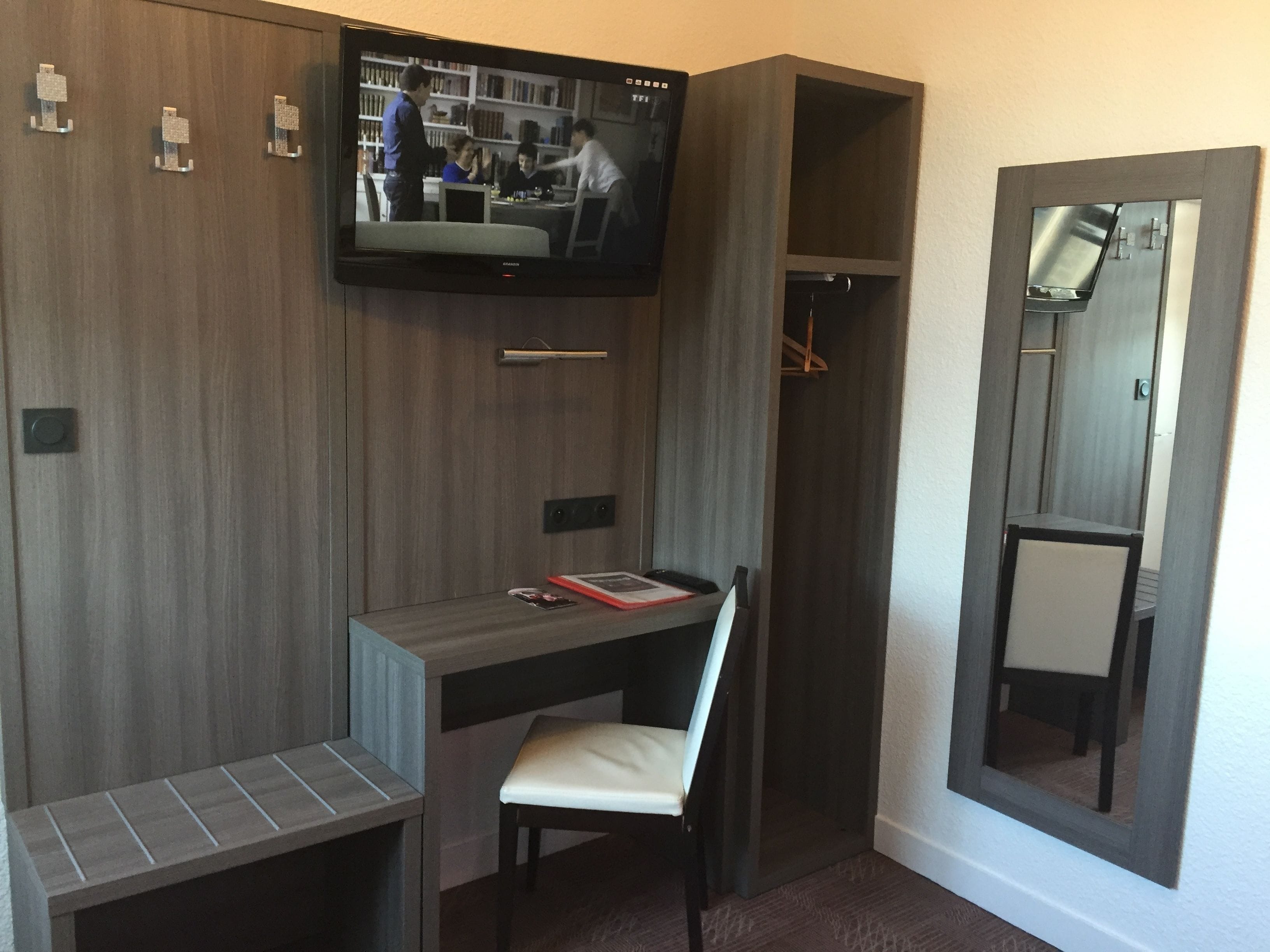 A hotel room with a built-in desk, a television mounted on the wall, a mirror, and a wardrobe.