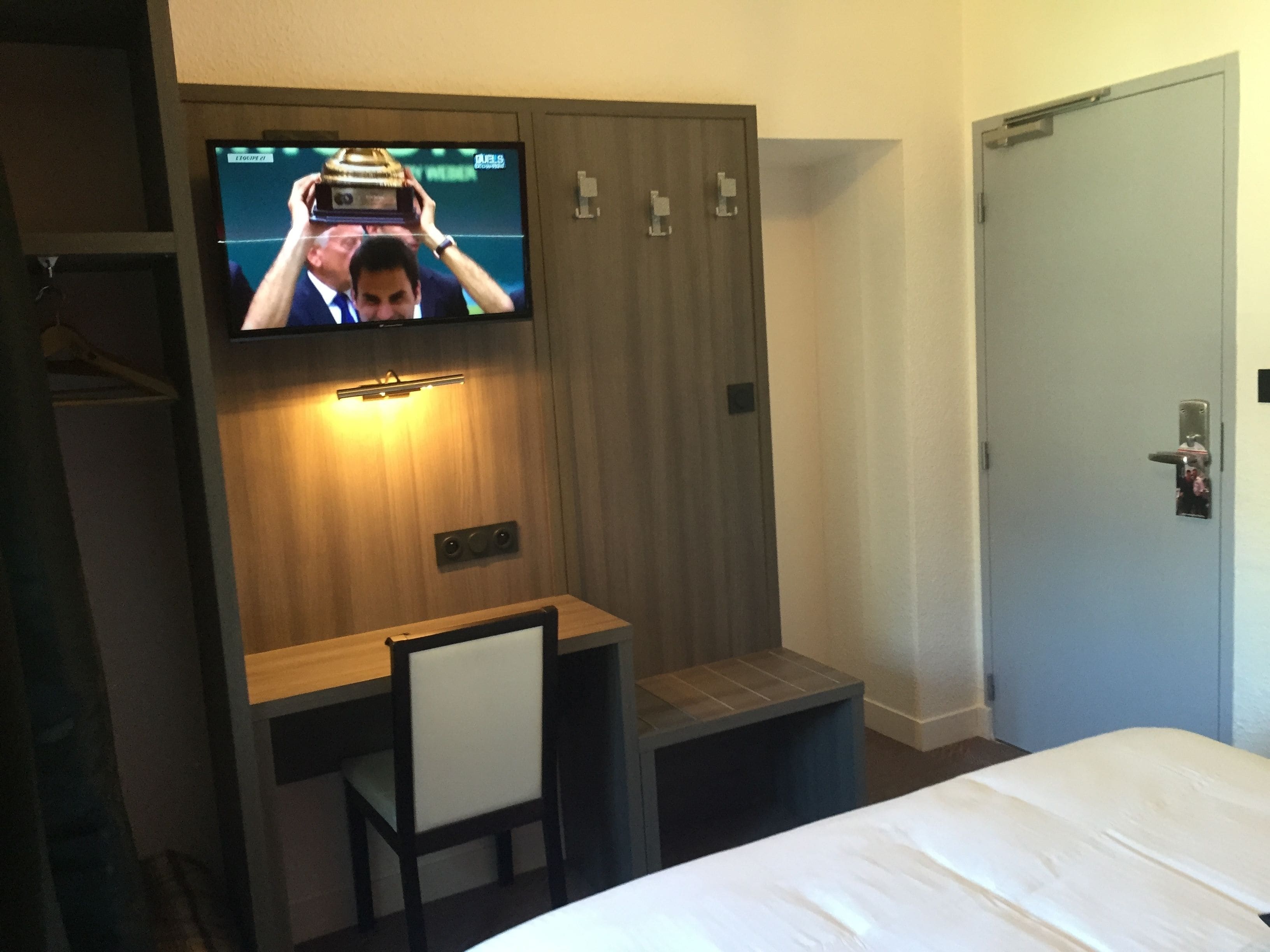 Una habitación de hotel con cama, escritorio, silla, televisor montado en la pared y una puerta.