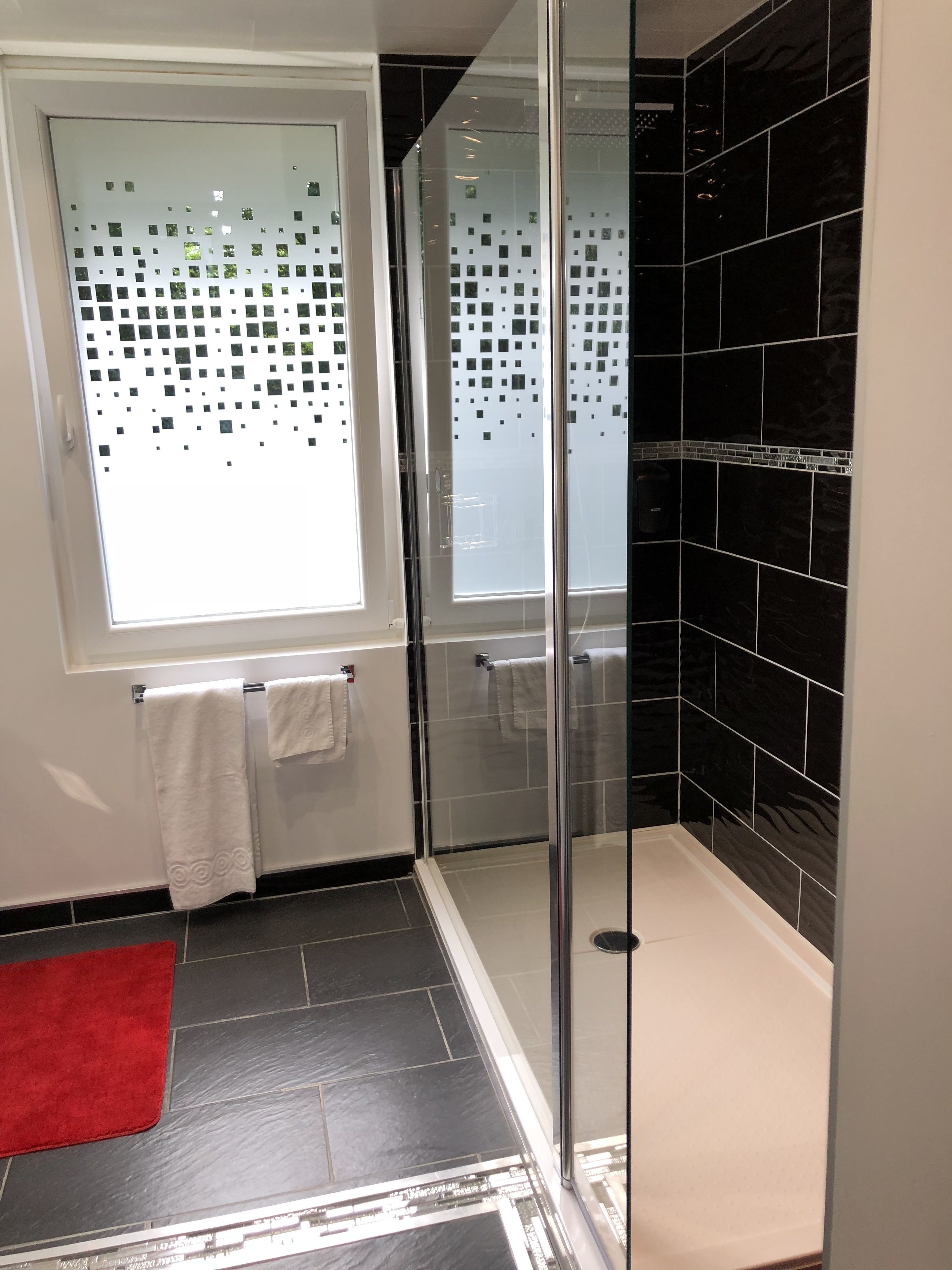 Un baño moderno con cabina de ducha de vidrio, una ventana con vidrio esmerilado y paredes cubiertas de azulejos negros.