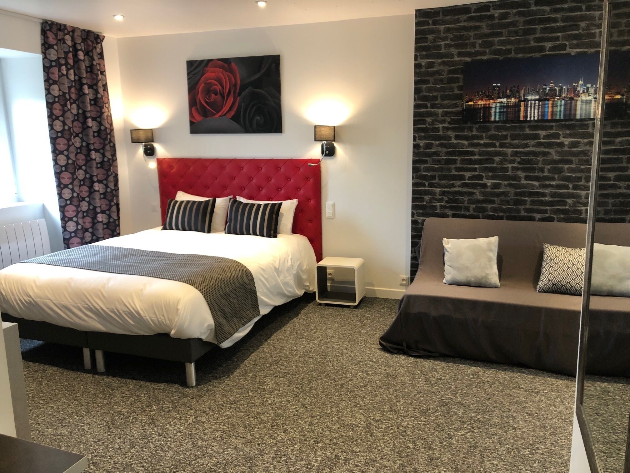 A hotel room with a red tufted headboard, a bed with striped pillows, a grey sofa with patterned cushions, a cityscape wall art, and a small table lamp.