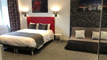 A hotel room with a red tufted headboard, a bed with striped pillows, a grey sofa with patterned cushions, a cityscape wall art, and a small table lamp.