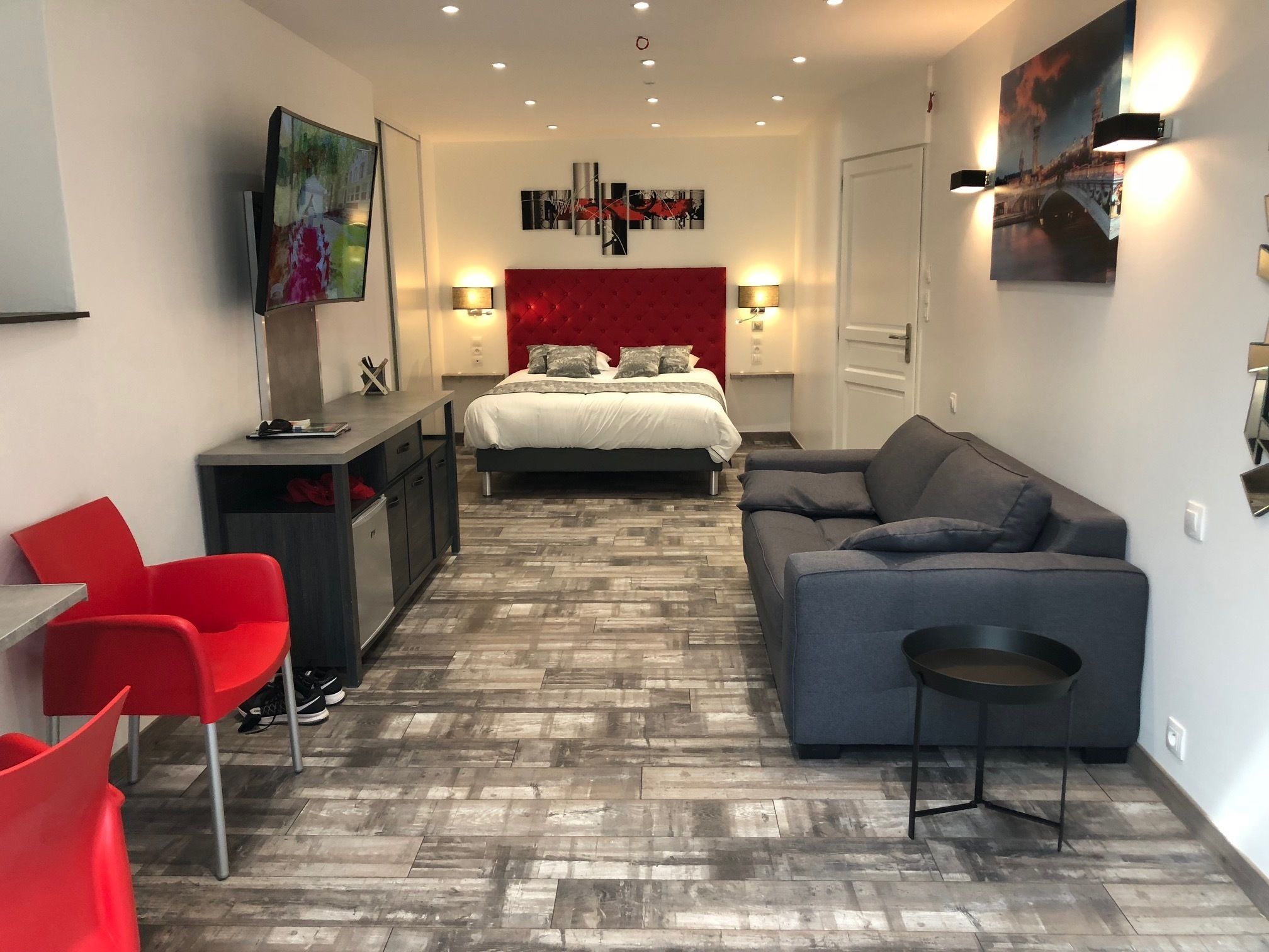 Une chambre d’hôtel moderne avec un lit, une tête de lit rouge, un canapé gris, une chaise rouge, une petite table et un téléviseur.