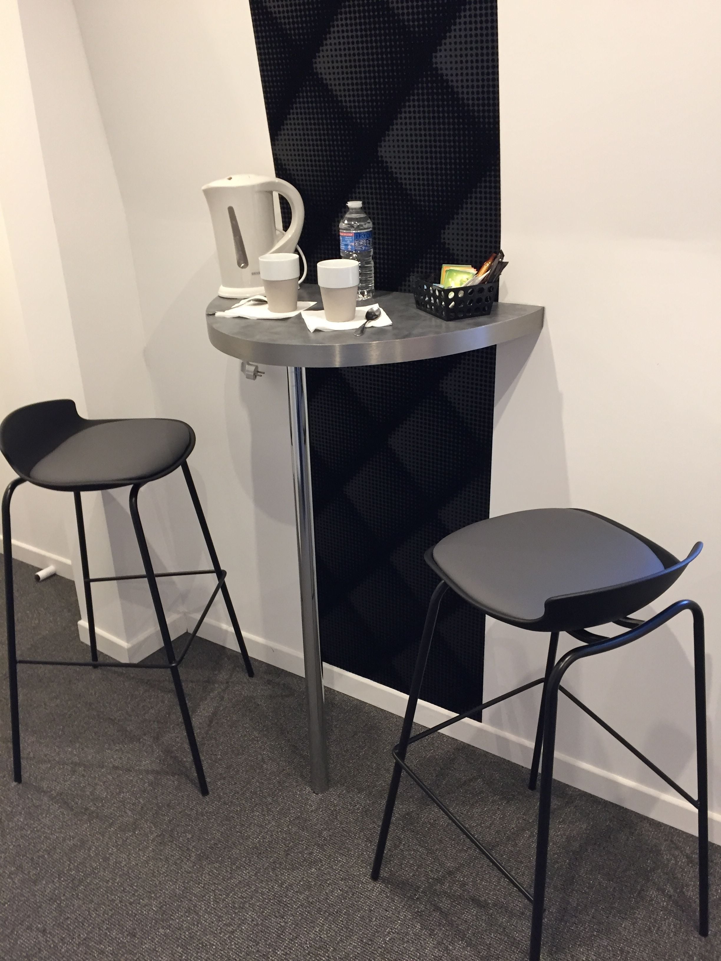 A small, modern room with two black bar stools, a small table with a water bottle, a coffee maker, and a basket of keys.