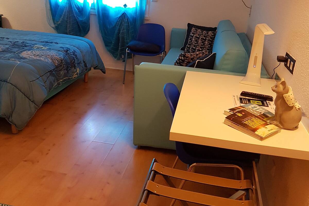 A room with a bed, a desk with a lamp, a chair, a sofa, and a small table with books and a figurine.