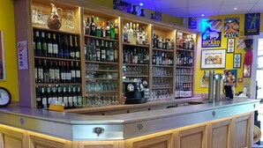 Una zona de bar con una barra central, estanterías con botellas y vasos, y un taburete en primer plano.