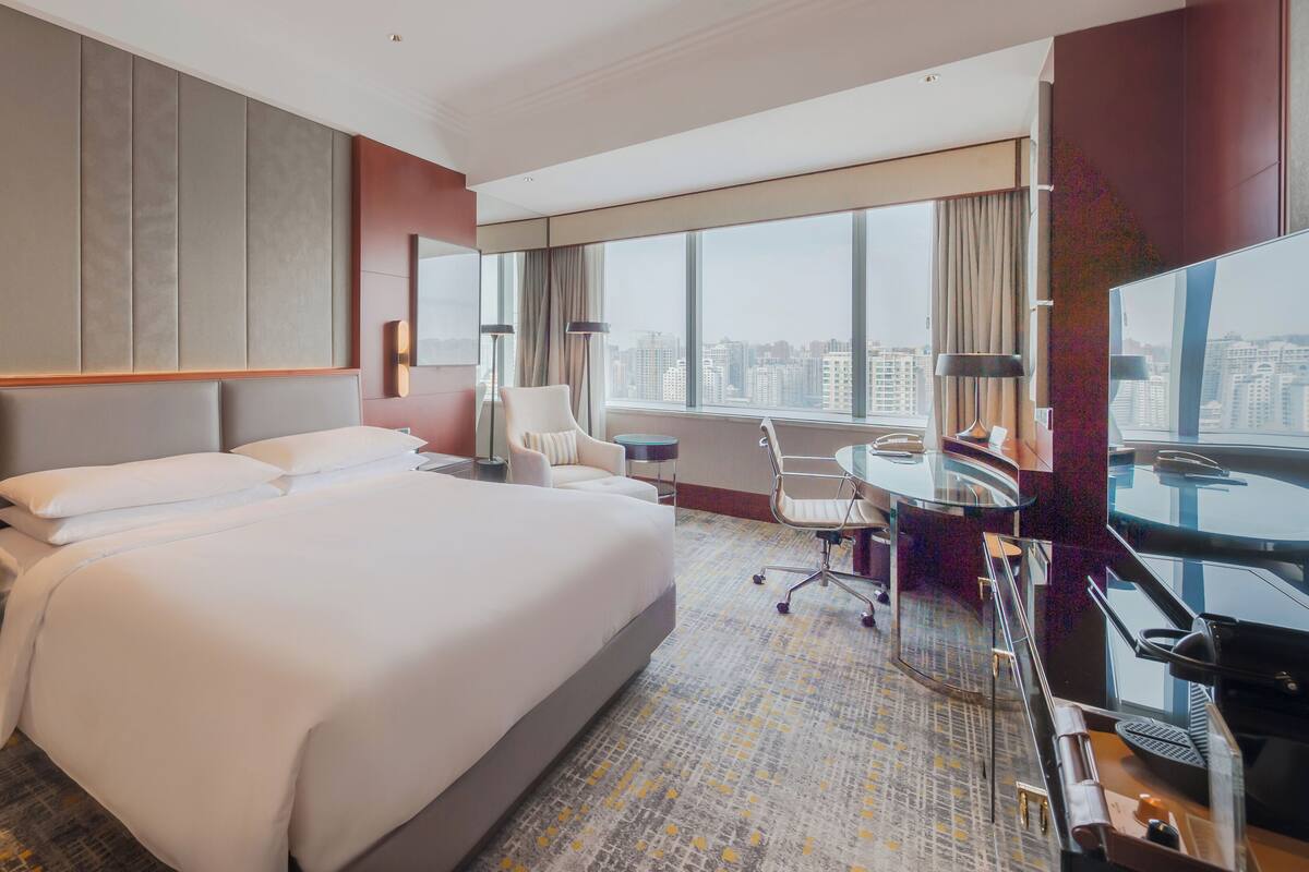 Habitación de hotel con una cama grande, un escritorio, una silla, un televisor y vistas a la ciudad.