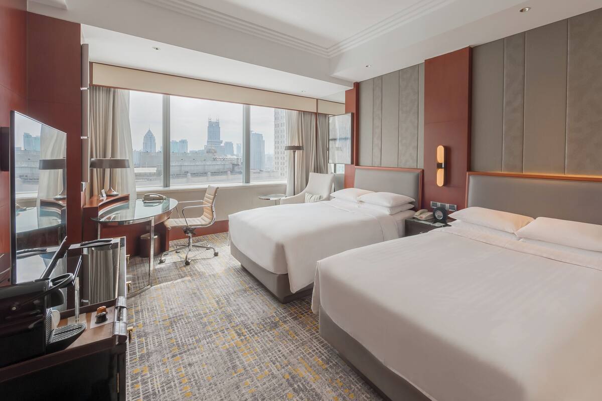 Habitación de hotel con dos camas, un escritorio, una silla, una lámpara de escritorio y vistas a la ciudad.