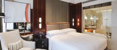 A modern hotel room with a large bed, a seating area with a chair and a small table, a large mirror, and a bathroom with a bathtub and a sink.
