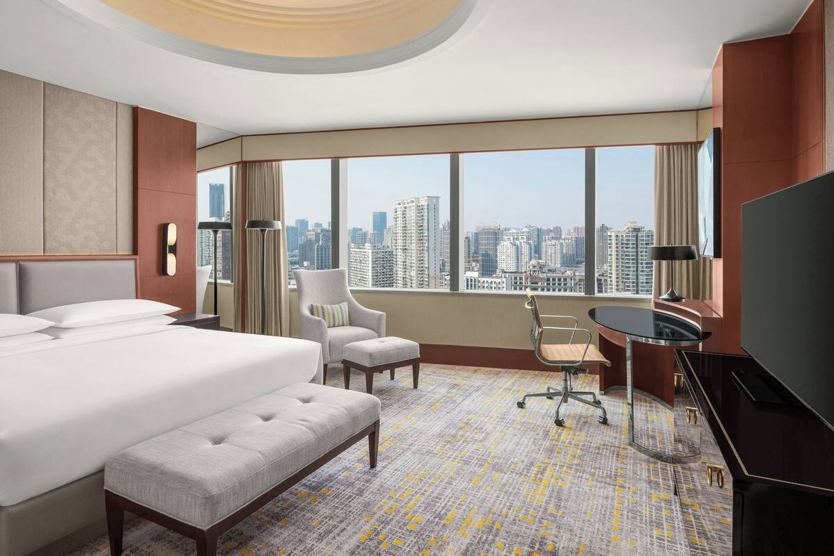 Habitación de hotel con una cama grande, un escritorio, una silla y un televisor. Cuenta con un ventanal con vistas a la ciudad.