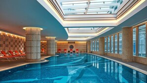 Una piscina interior moderna con techo de tragaluces, sillones reclinables rojos y pilares de piedra.