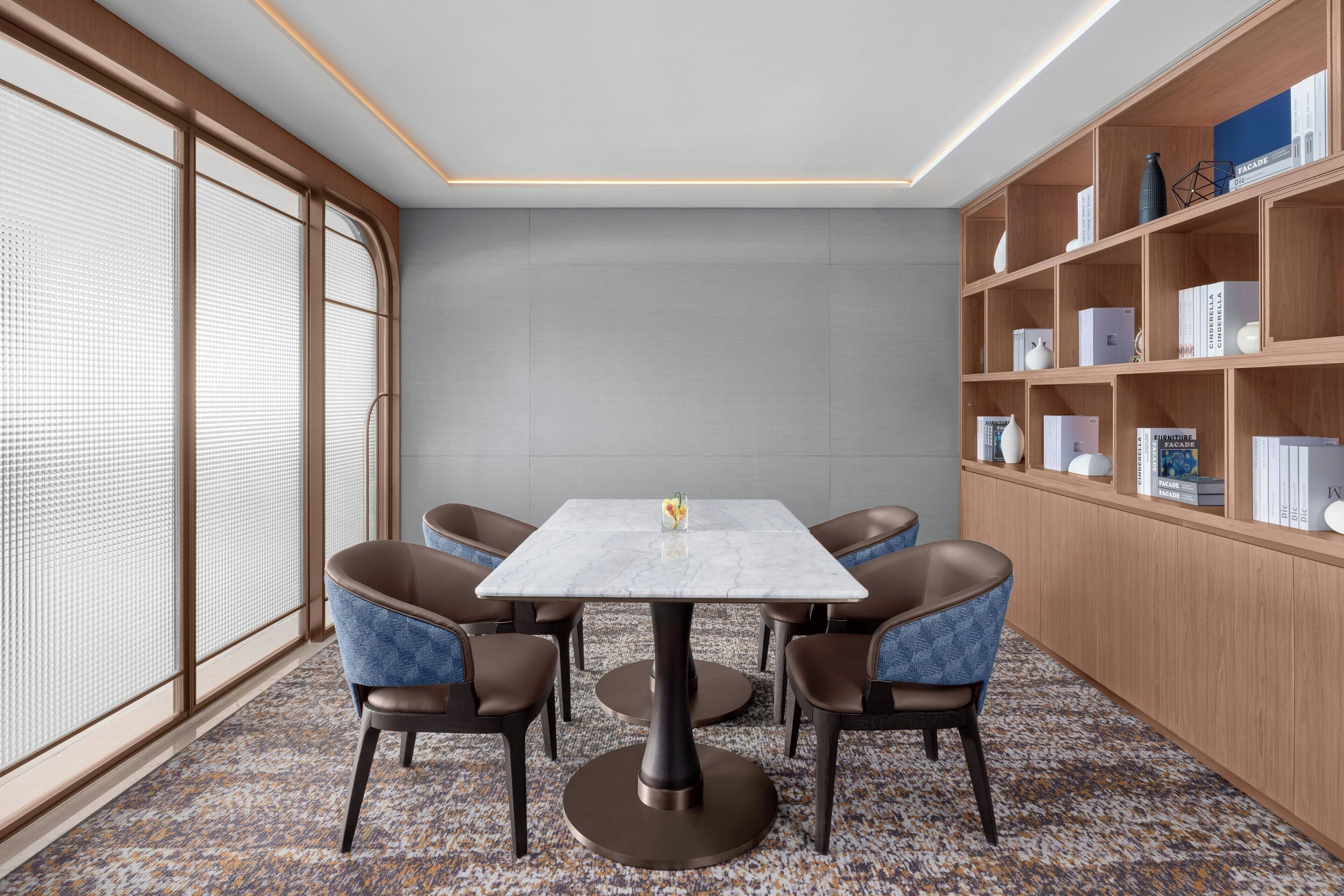 A modern conference room with a marble table, four upholstered chairs, a bookshelf with books and decorative items, and large windows with blinds.