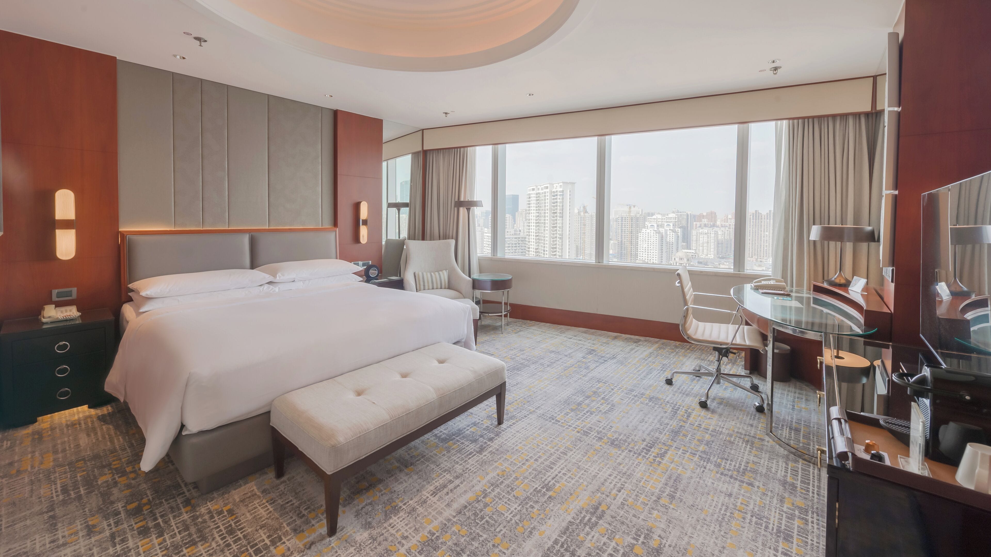 A hotel room with a large bed, a desk, a chair, a bench, and a view of the cityscape.