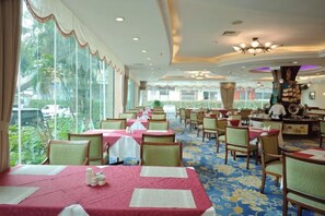 Ein Speisesaal eines Hotels mit gedeckten Tischen, großen Fenstern mit Blick ins Freie und einer Decke mit Einbaustrahlern.