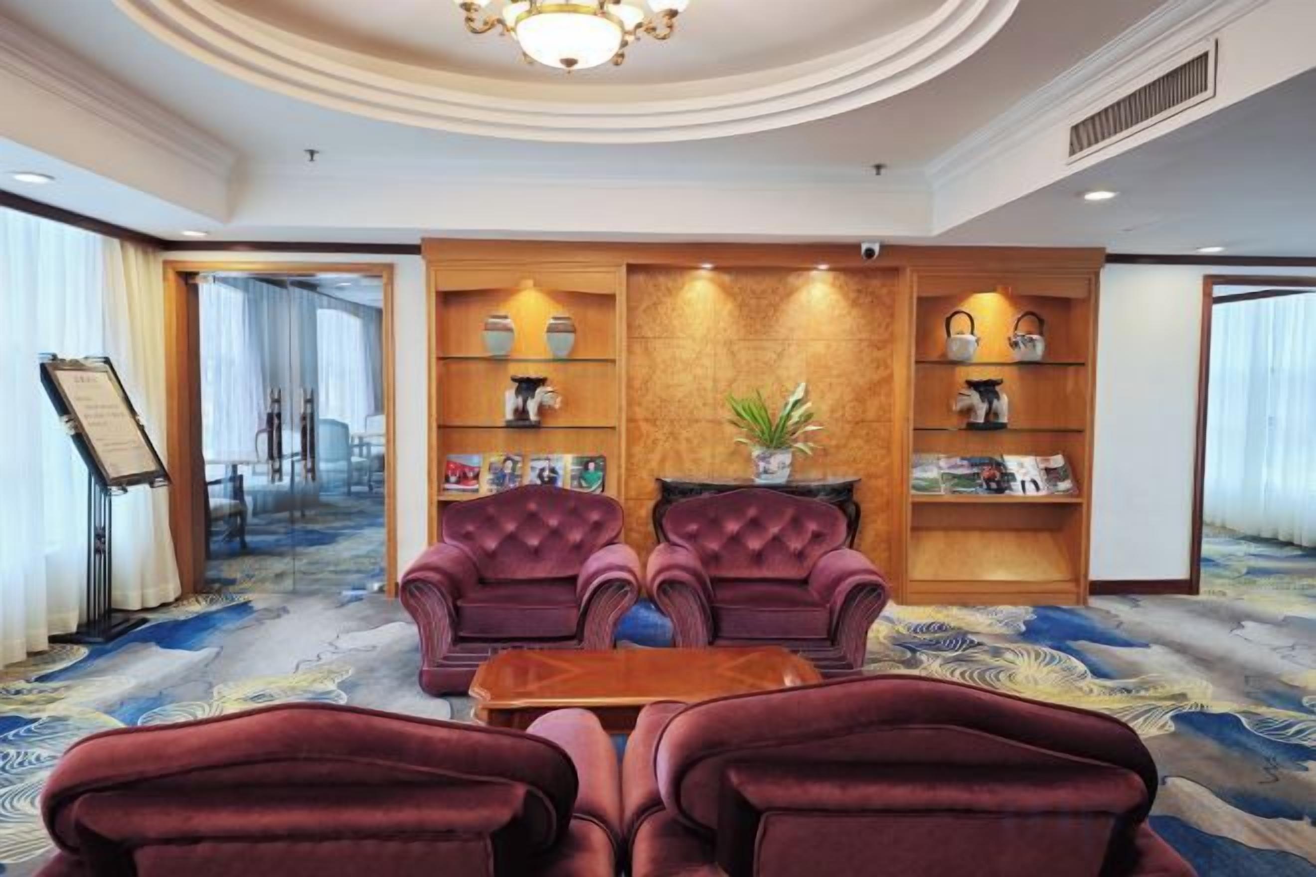 Een hotellobby met een vloerbedekking, twee fauteuils, een houten bijzettafeltje en een boekenkast met decoratieve objecten.