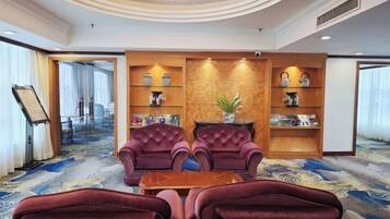 Een hotellobby met een vloerbedekking, twee fauteuils, een houten bijzettafeltje en een boekenkast met decoratieve objecten.