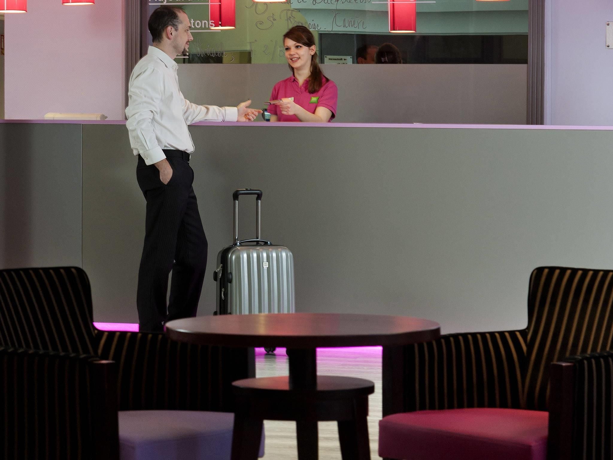 Een hotel receptie met een balie, een zittende gast en een staande gast.