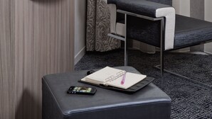 Habitación de hotel moderna con televisor de pantalla plana montado en la pared, un estante blanco pequeño con una taza, un kiwi y una naranja, una silla tapizada de color gris, y un reposapiés negro con un cuaderno y un bolígrafo.