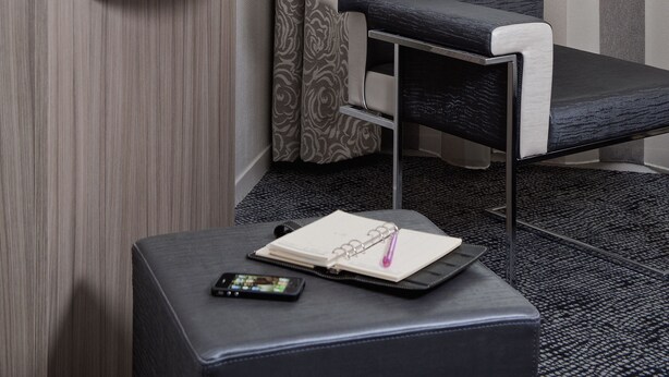 A modern hotel room with a flat-screen TV mounted on the wall, a small white shelf with a cup, a kiwi, and an orange, a grey upholstered chair, and a black ottoman with a notebook and pen.