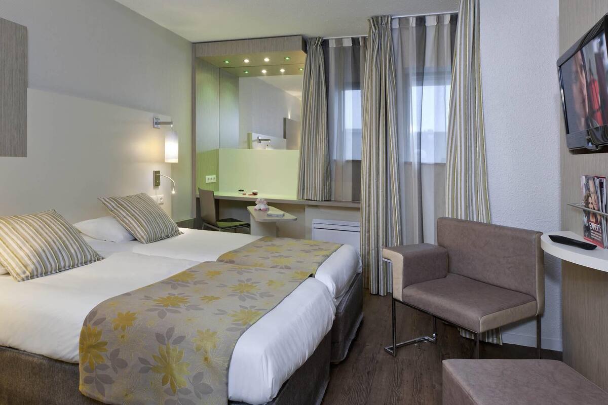Quarto de hotel com cama, escrivaninha, cadeira e televisão.