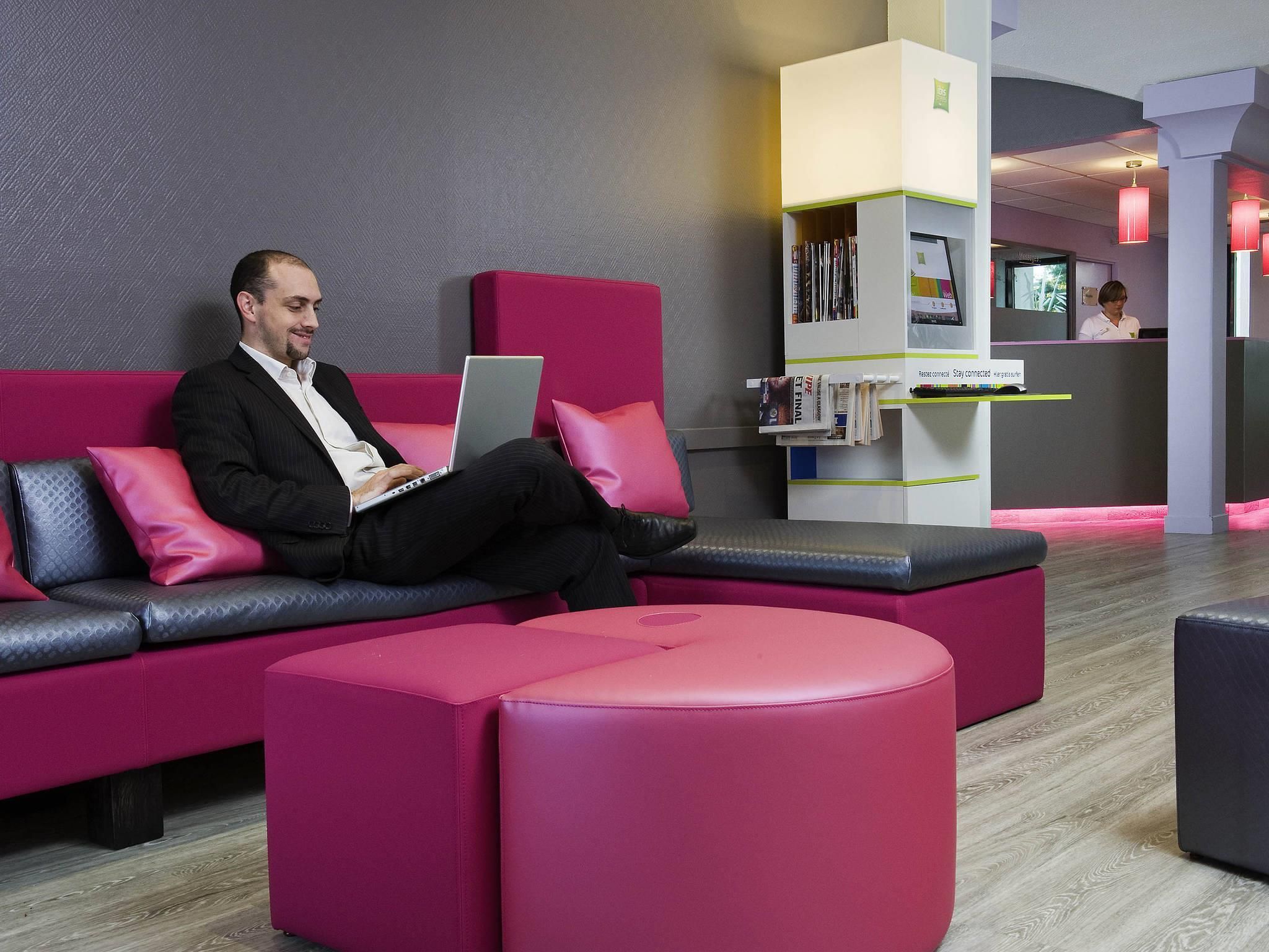A man sitting on a pink sofa with a laptop, a pink ottoman, and a reception desk with magazines and a TV in the background.
