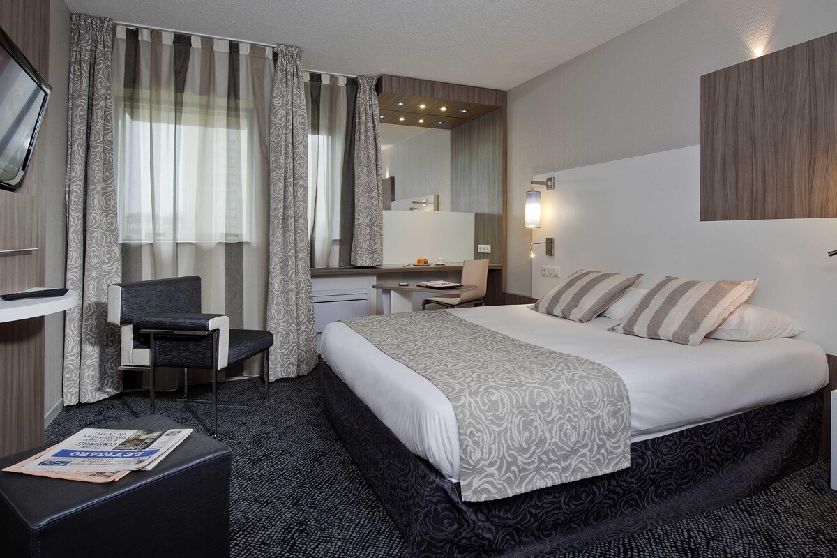 Una habitación de hotel con cama, escritorio, silla, televisión y espejo de baño.