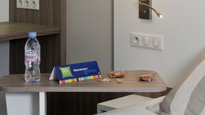 A hotel room with a bed, a wooden nightstand, a water bottle, a welcome card, and a wall-mounted light fixture. - ibis Styles Melun Hotel (Rubelles)
