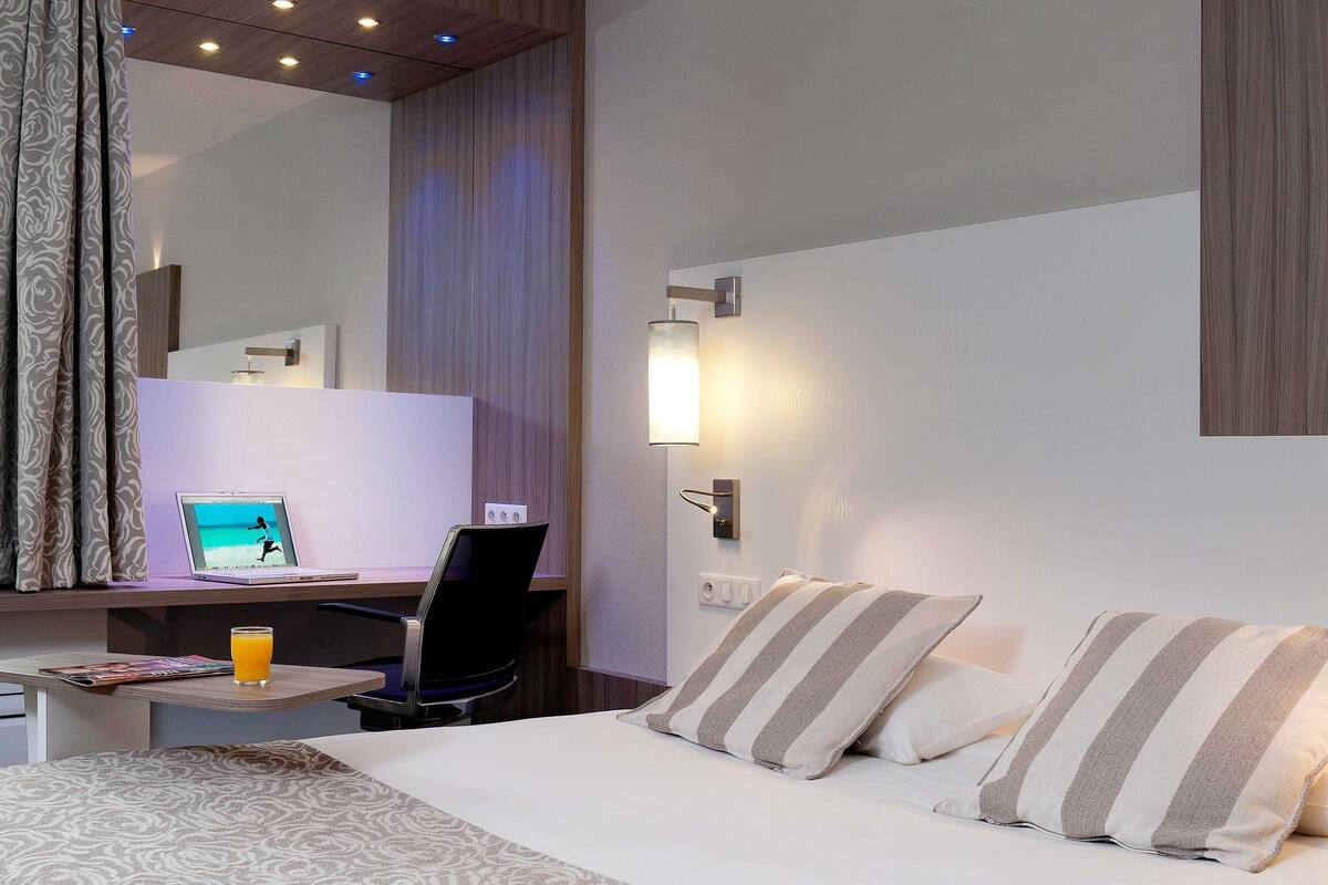 Quarto de hotel com cama, escrivaninha com laptop, cadeira, mesa de cabeceira com copo de suco de laranja e luminária na parede.