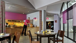 A modern hotel dining area with wooden furniture, a kitchen in the background, and large windows.
