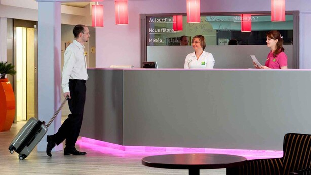 Zona de recepción de hotel con diseño moderno, que incluye un mostrador de recepción, un área de espera con una mesa redonda y sillas, y un ascensor visible al fondo.