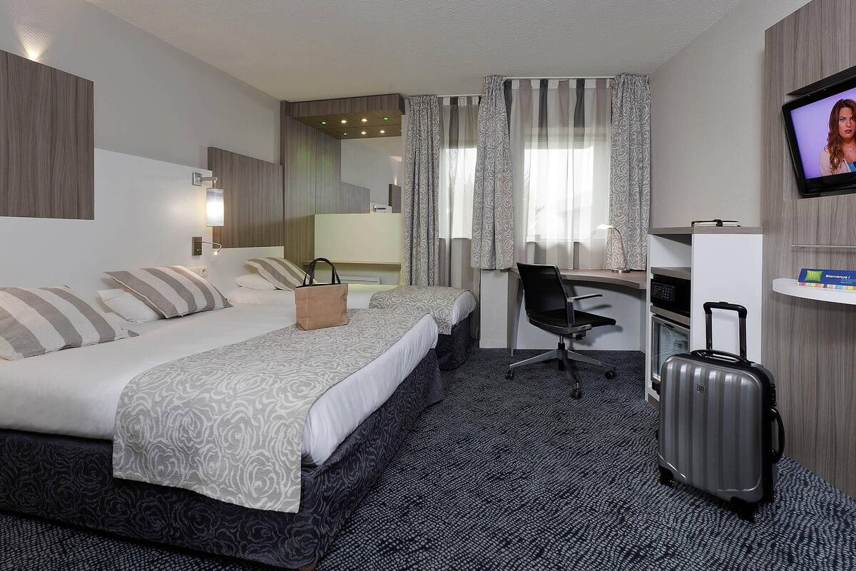 Quarto de hotel com duas camas, uma escrivaninha, uma cadeira, uma televisão e uma mala.