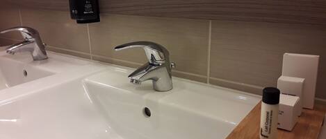 A bathroom with two sinks, a mirror, and a wooden shelf with a shoe sponge and a bottle.