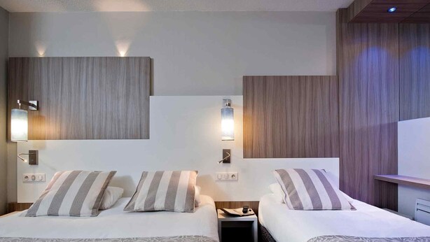 Habitación de hotel con dos camas, una mesita de noche, iluminación empotrada en la pared y un panel de pared de madera.