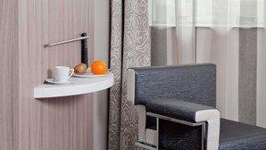 Habitación de hotel con pared de madera, televisor montado, repisa blanca con una taza, un kiwi y una naranja, y una silla tapizada de color gris.