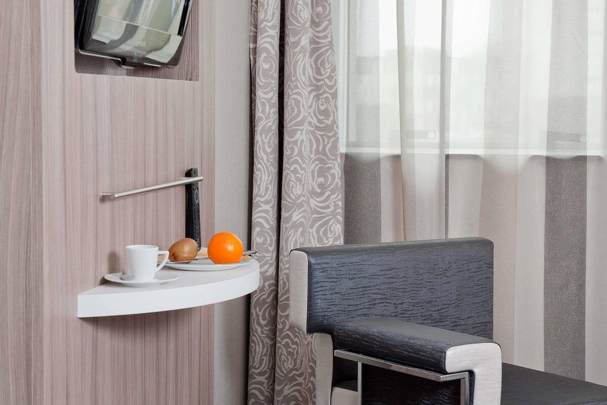Quarto de hotel com parede de madeira, televisão montada, prateleira branca com uma xícara, um kiwi e uma laranja, e uma cadeira estofada cinza.